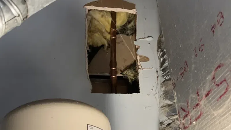 Hole in wall revealing insulation and copper pipe inside a utility room with exposed ductwork and tank.