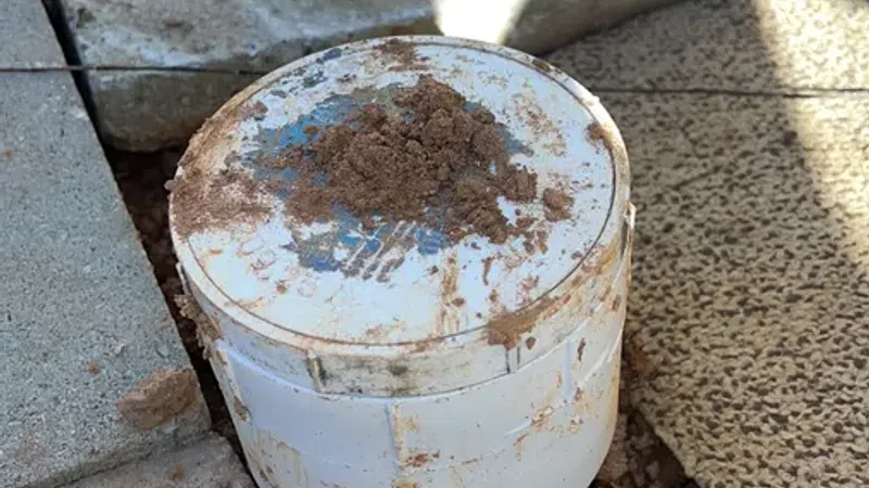 Close-up of a white PVC pipe cap with dirt on top, surrounded by concrete tiles and garden hoses outdoors.