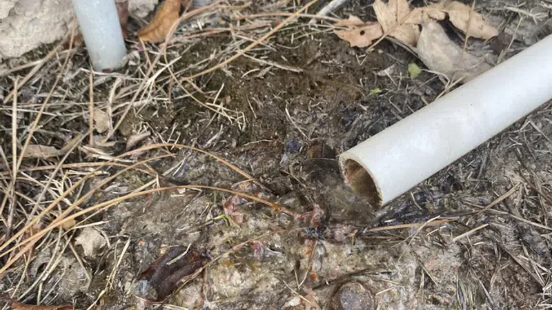 Outdoor ground with dirty water draining from a broken white PVC pipe near a stucco wall with dry leaves and debris.