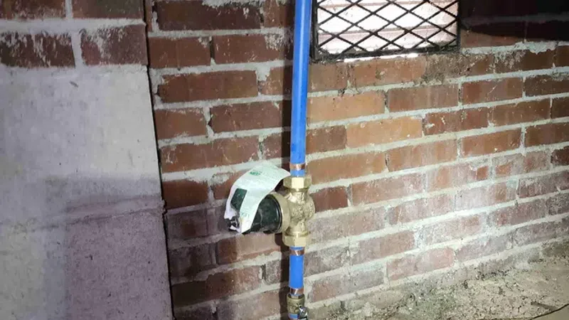 Blue water pipes connected to a meter installed in a basement with brick walls and concrete floor.