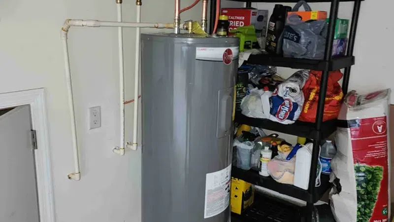 Gray cylindrical water heater installed on a metal drip pan near storage shelves with various items in a utility room.