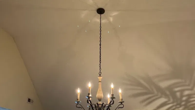 Lit chandelier hanging from high ceiling casting shadows in cozy living room with fireplace and windows.
