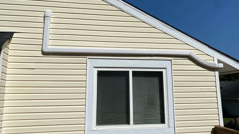 Beige house wall with white framed window and exterior white PVC piping against clear blue sky.