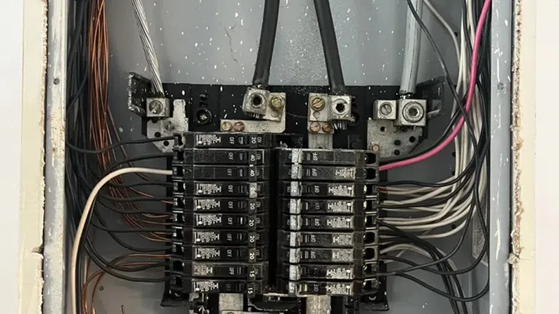 Open electrical panel with circuit breakers and multiple black, white, and copper wires inside a gray metal box.