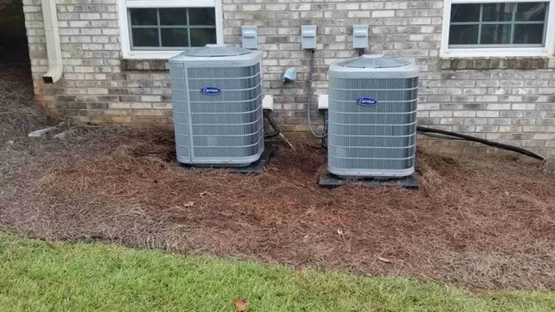 Two gray Carrier air conditioning units installed outside a brick house between two windows on a mulch bed.