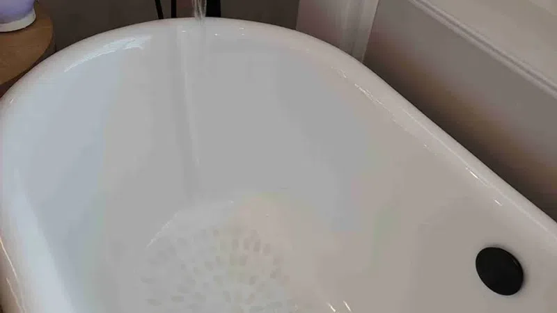 White freestanding bathtub with black faucet and running water next to a window in a bathroom.