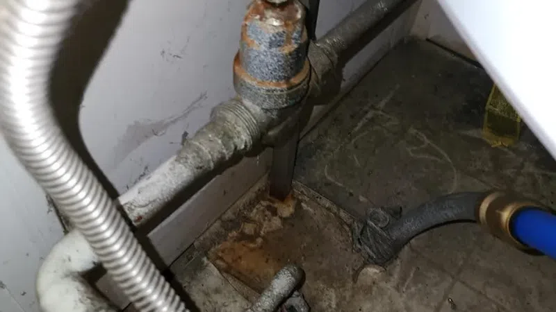 Rusty and corroded plumbing pipes and valves under a sink with visible dirt and grime on the floor.
