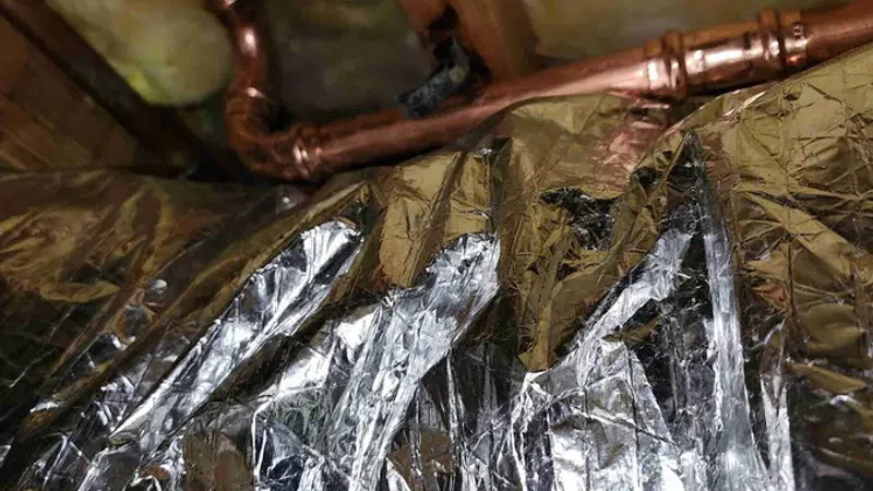 Close-up of copper pipes and yellow insulation with reflective foil radiant barrier in attic space.