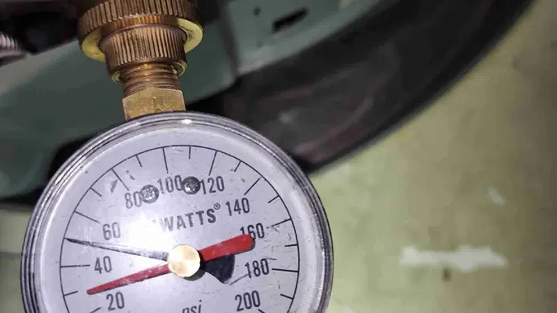 Close-up of a Watts pressure gauge reading around 160 psi connected to a brass valve on a water heater.