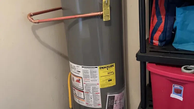 Gray Rheem water heater installed in a corner with copper pipes and shelving with storage bins nearby.