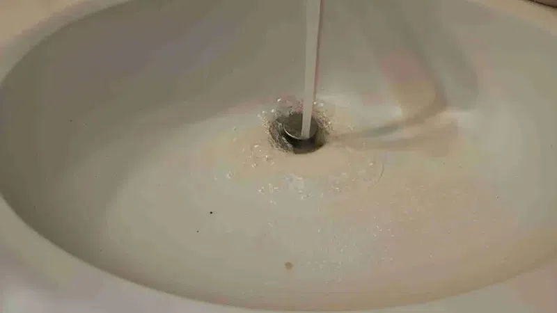 Running water flowing from a chrome faucet into a white bathroom sink with soap and toiletries on the side.