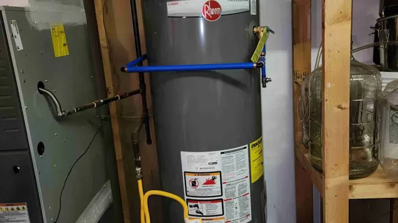 Gray Rheem water heater installed in a basement with pipes, ductwork, and wooden shelving nearby.