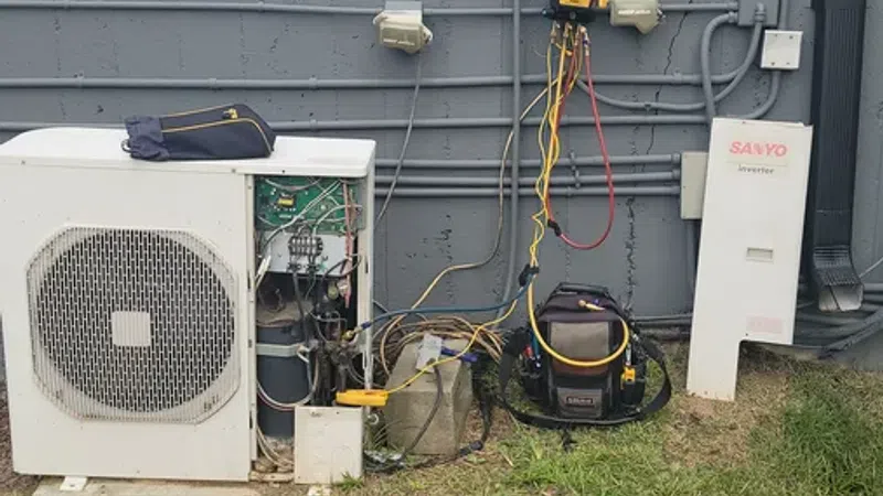 Outdoor air conditioning unit with open panel, diagnostic tools, and wiring arranged against a gray wall with grass foreground.