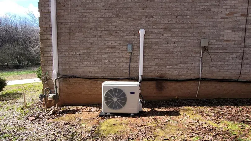 HVAC outdoor unit installed on ground beside a brick house wall with fallen leaves covering the yard.