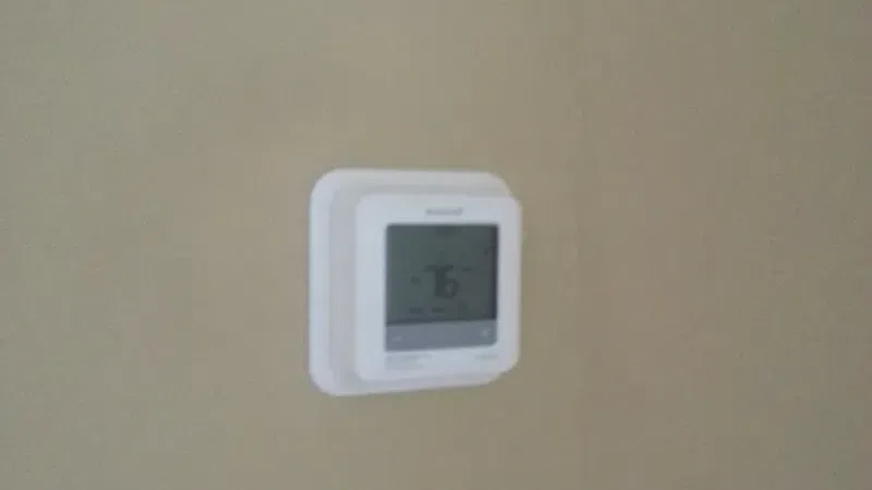 Wall-mounted white thermostat displaying temperature set to 76 degrees on beige wall.