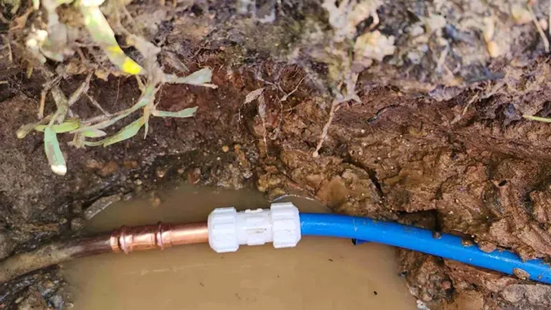Water pipe connection between copper and blue plastic pipes submerged in muddy soil with surrounding grass.