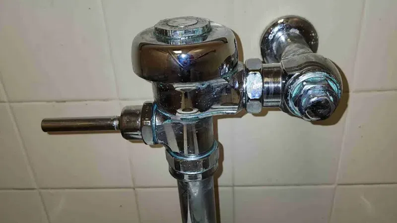 Close-up of a chrome flush valve assembly attached to a white toilet tank against tiled wall.
