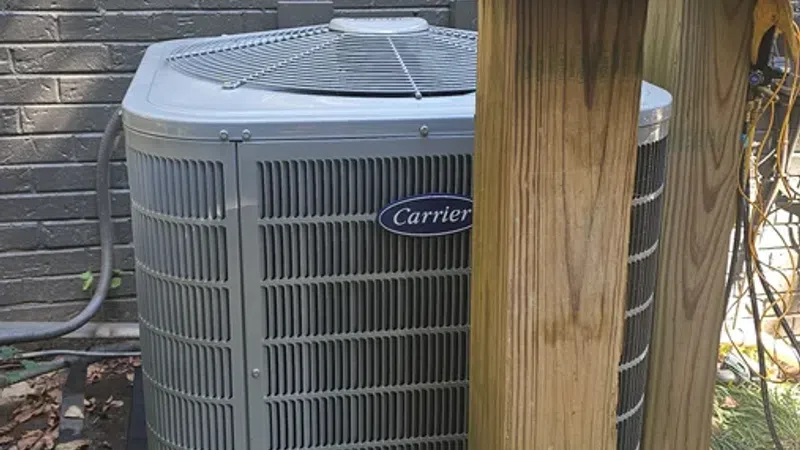 Carrier brand air conditioning unit installed outdoors on a concrete slab next to a brick wall and wooden posts.