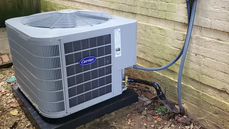 Outdoor Carrier air conditioning unit installed on a black base outside a house with brick walls and gravel ground.