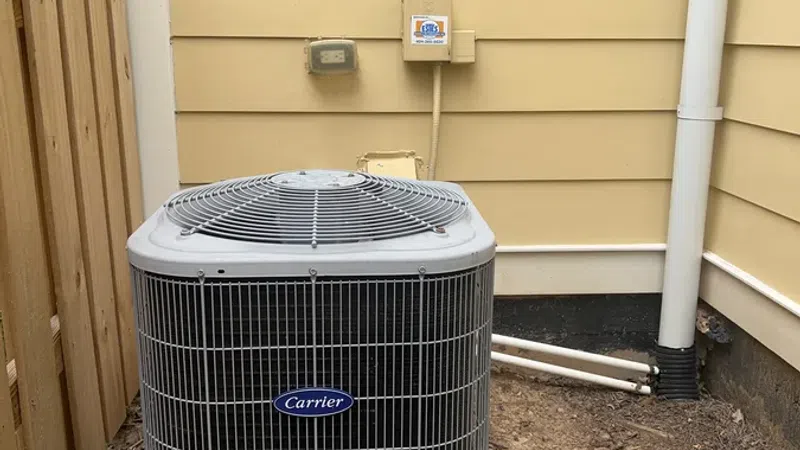 Carrier outdoor air conditioning unit installed on concrete slab beside yellow house with wooden fence.