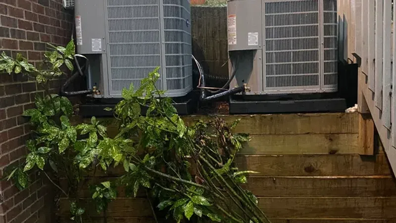 Two outdoor HVAC units placed on a raised wooden platform beside a brick wall and a staircase with plants and wet rocks below.