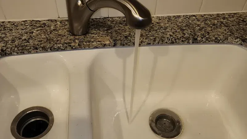 Modern stainless steel faucet running water into a white double kitchen sink with granite countertop.