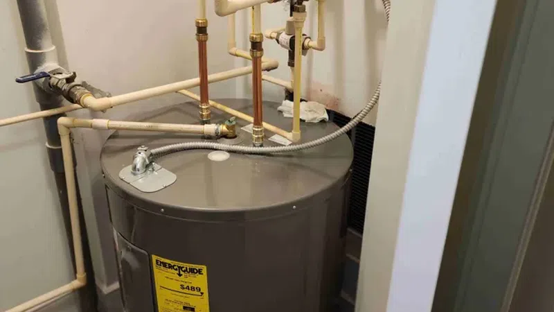 Residential water heater tank with connected copper and PVC pipes and pressure gauge in utility room.