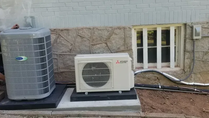 Two outdoor air conditioning units installed beside a white brick house with a small basement window.