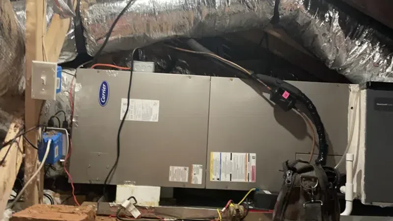 HVAC unit installed in attic with insulated ductwork and tool bag nearby on wooden floor under roof beams