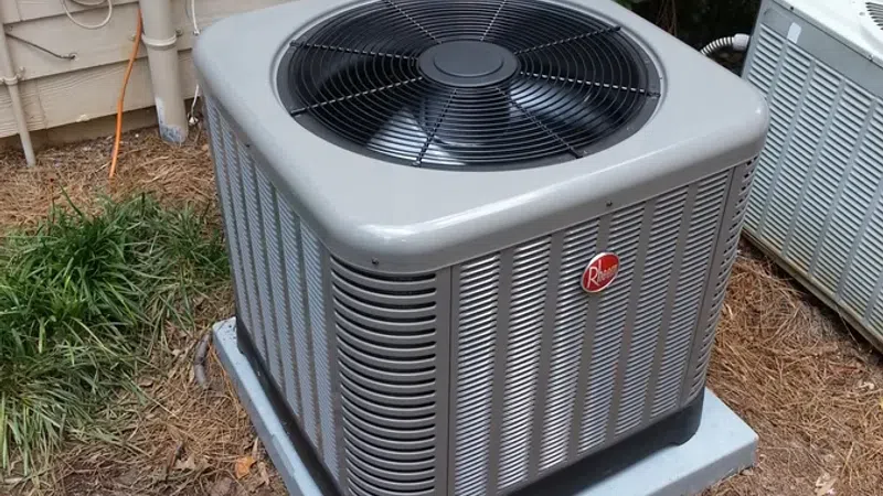 Outdoor Rheem air conditioning unit installed on a concrete pad beside a house with greenery nearby.