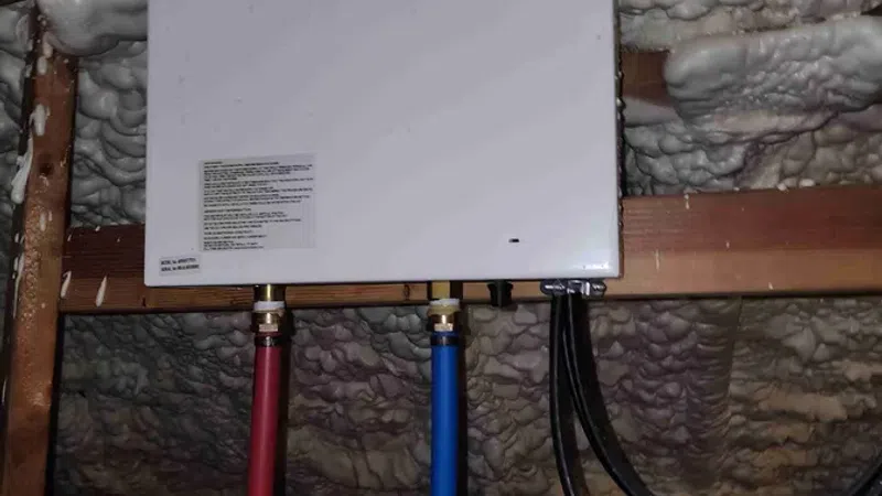 White tankless water heater mounted in attic with red and blue water pipes and foam insulation around