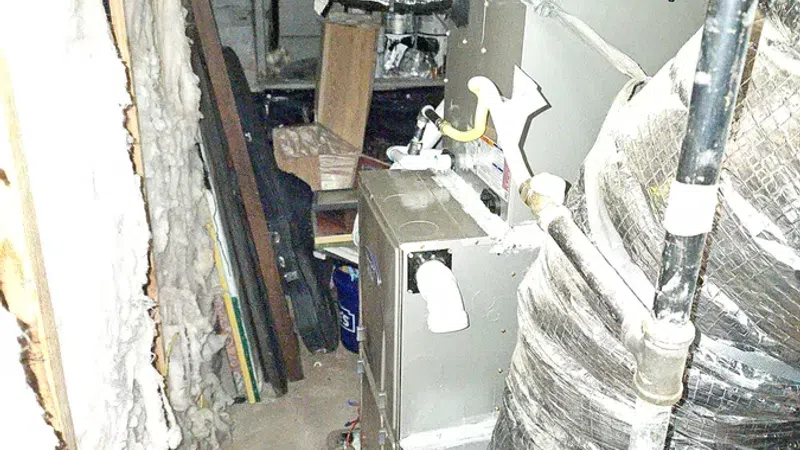 Narrow basement area with HVAC system, ductwork wrapped in insulation, pipes, and cluttered storage space.