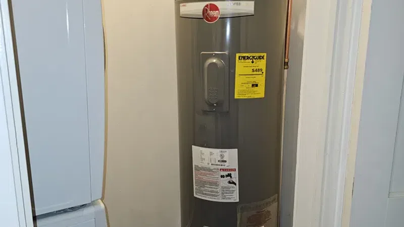 Gray Rheem water heater installed in a small room with copper pipes and energy guide label visible.