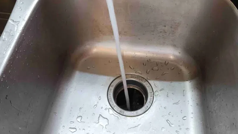 Stream of water flowing from faucet into a wet stainless steel kitchen sink with drain strainer.