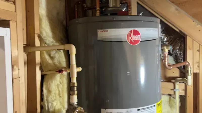 Gray Rheem Classic water heater installed in an attic with exposed insulation and wooden framing.