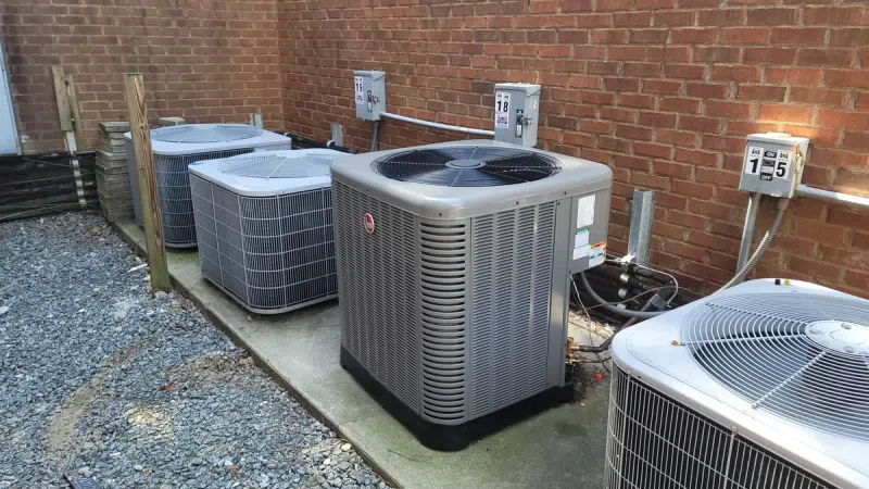 Row of outdoor HVAC air conditioning units installed beside a brick wall on concrete pads.