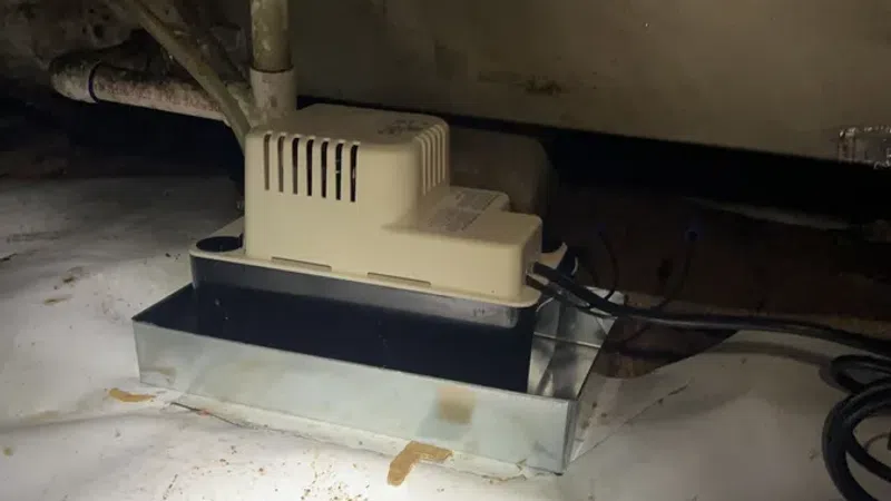 Condensate pump installed on a dirty basement floor with connected pipes and electrical wiring.
