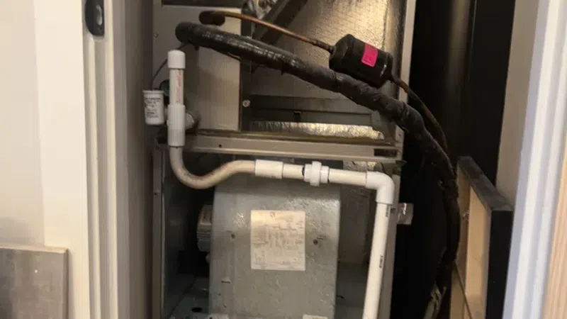 Open HVAC unit with exposed wiring, pipes, and metal heat exchanger in a small utility closet.