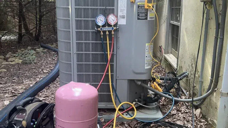 Outdoor HVAC unit with refrigerant gauges, pink refrigerant tank, and tools setup for maintenance near house wall.