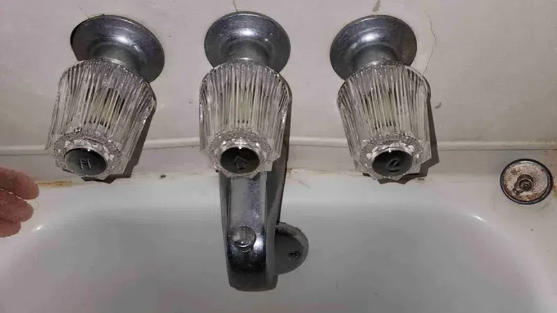 Old bathroom sink with three vintage glass faucet knobs labeled hot, water, and cold, showing wear.