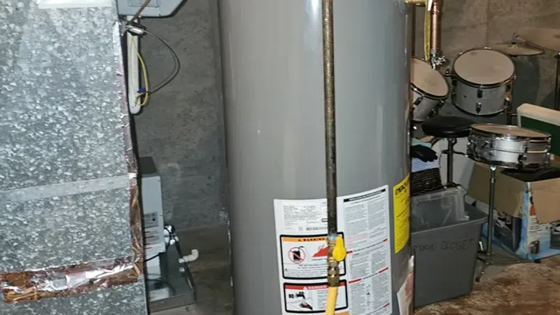 Gray water heater with copper pipes and a yellow gas line installed in a basement near ductwork and musical drums.