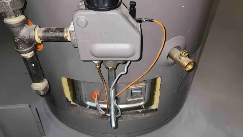 Close-up of a gas water heater control valve with connected pipes and warning labels on tank.