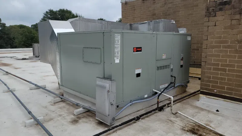 Large gray rooftop HVAC unit installed beside a brick wall on a flat roof under cloudy sky.