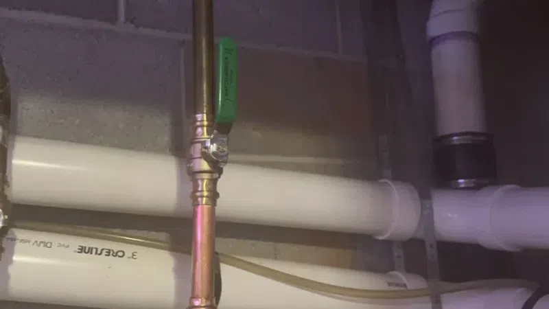 Copper and white PVC pipes connected with valves in a basement wall near insulation and ceiling wiring.