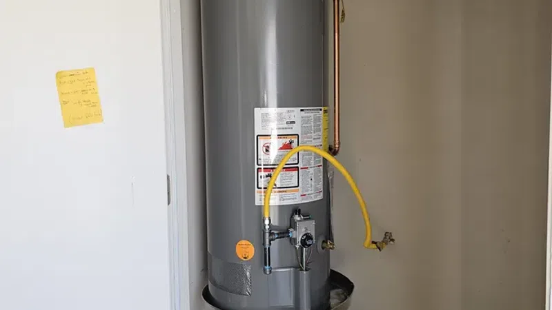 Tall gray Rheem water heater with yellow gas line installed in a small utility closet with concrete floor.