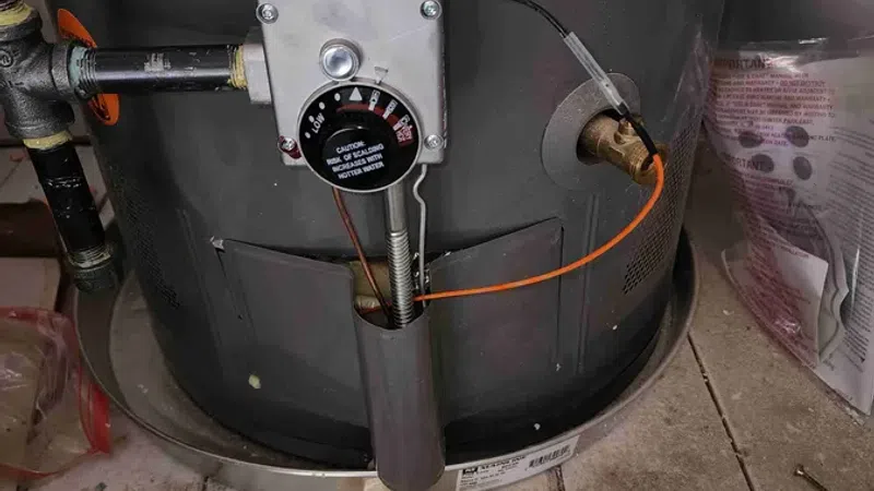 Close-up of a water heater control panel with temperature dial and connected pipes on tiled floor.
