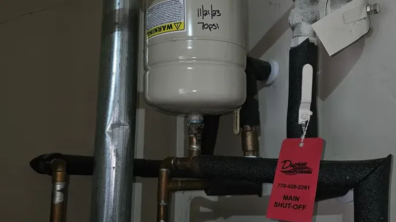 Water heater pipes and expansion tank with main shut-off valve and red tag in utility room corner.