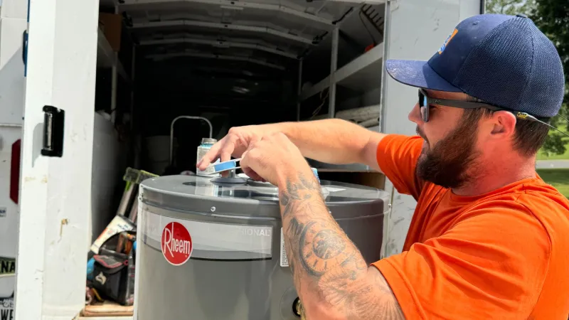 Estes Plumber in orange shirt and cap working on a replacement of Rheem water heater outdoors by a white service truck.