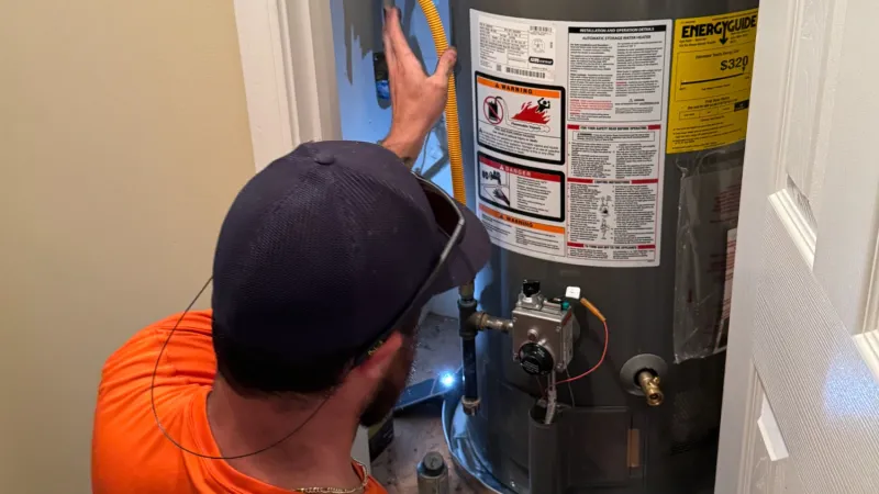 Estes Plumber in orange uniform inspecting and repairing a large gray water heater with yellow hose indoors.
