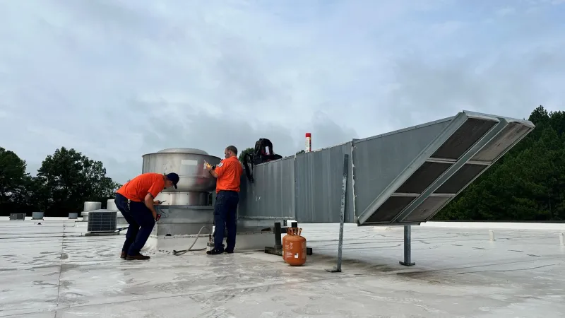 Two technicians in orange shirts inspecting rooftop HVAC equipment under a cloudy sky with trees in the background